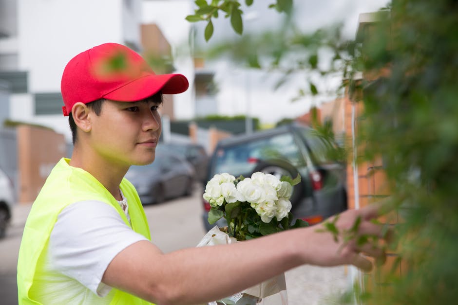 A young male flower delivery courier dressed in a neon yellow safety vest and a red cap is shown in profile, carefully arranging a bouquet of white roses and hydrangeas wrapped in white paper. He is s