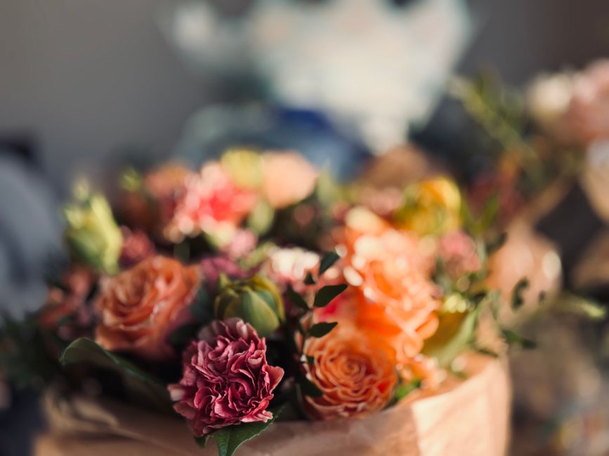 A close-up view of a floral arrangement featuring peach roses, deep pink carnations, and green foliage, arranged in a rustic, brown wrapping paper. The bouquet is placed on a surface with soft natural